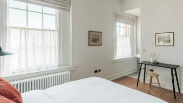 Une chambre avec un grand lit, deux fenêtres équipées de stores, un radiateur, un bureau avec une chaise et deux tableaux encadrés accrochés au mur.