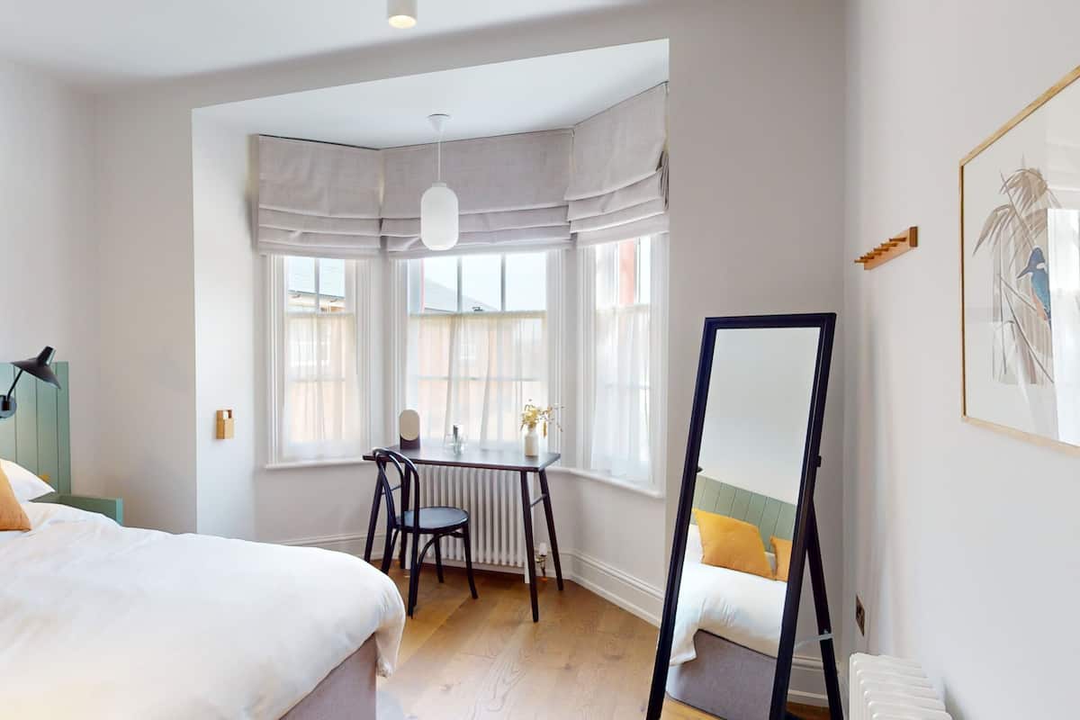 A bedroom with a bed, a desk, a chair, a mirror, and a window with curtains.
