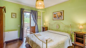 Un dormitorio con cama, mesita de noche, lámpara, ventana con cortinas y un cuadro en la pared.