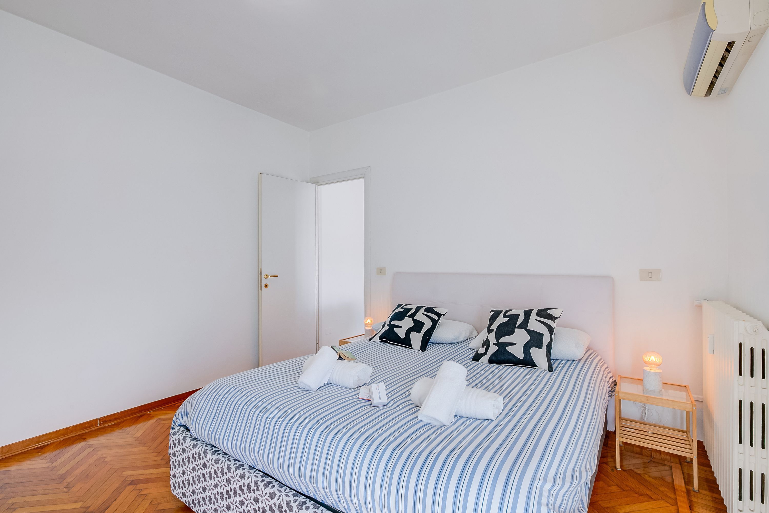 En pænt redt seng med stribet sengetøj, et natbord med lampe og en radiator.