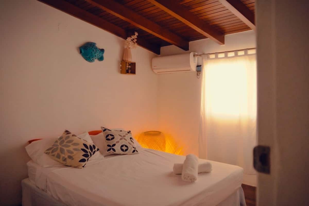 Ein Schlafzimmer mit einem Bett, Kissen, einem Nachttisch, einer Lampe und einem Fenster mit Vorhängen.