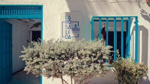A building with a blue door and window, a sign with 'LA ROSEA' and 'Los Rojos' text, and a white flowering tree in front. - Posada La Rosaleda (Los Roques)