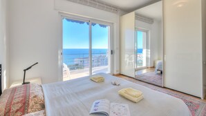 A bedroom with a large bed, a bedside table with a lamp, a book, and towels. There is a sliding glass door leading to a balcony with a view of the sea. - Sofy Apartment - Sea View (Sanremo)