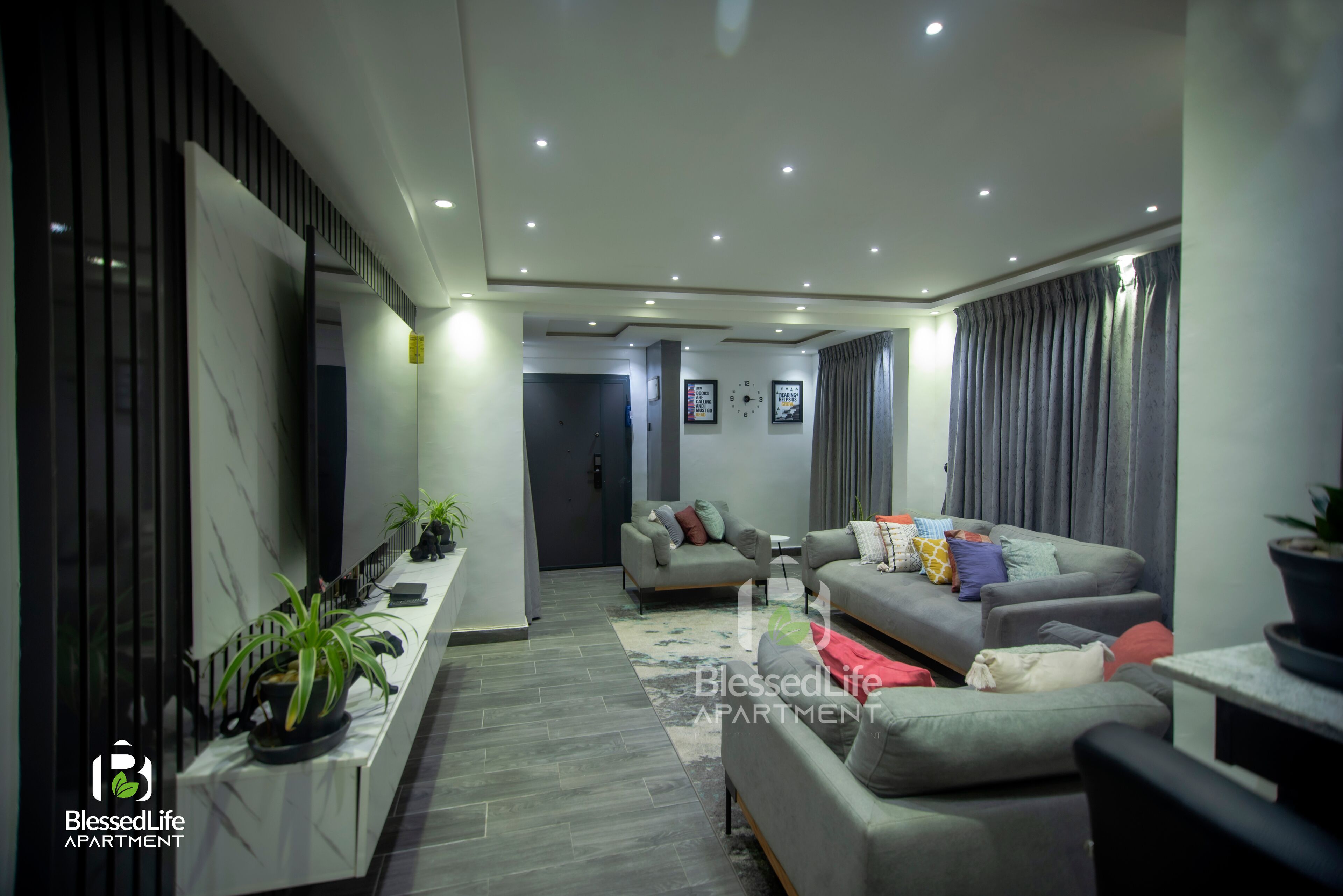 A modern living room with a sofa, a coffee table, and a TV. There are potted plants and a window with curtains.