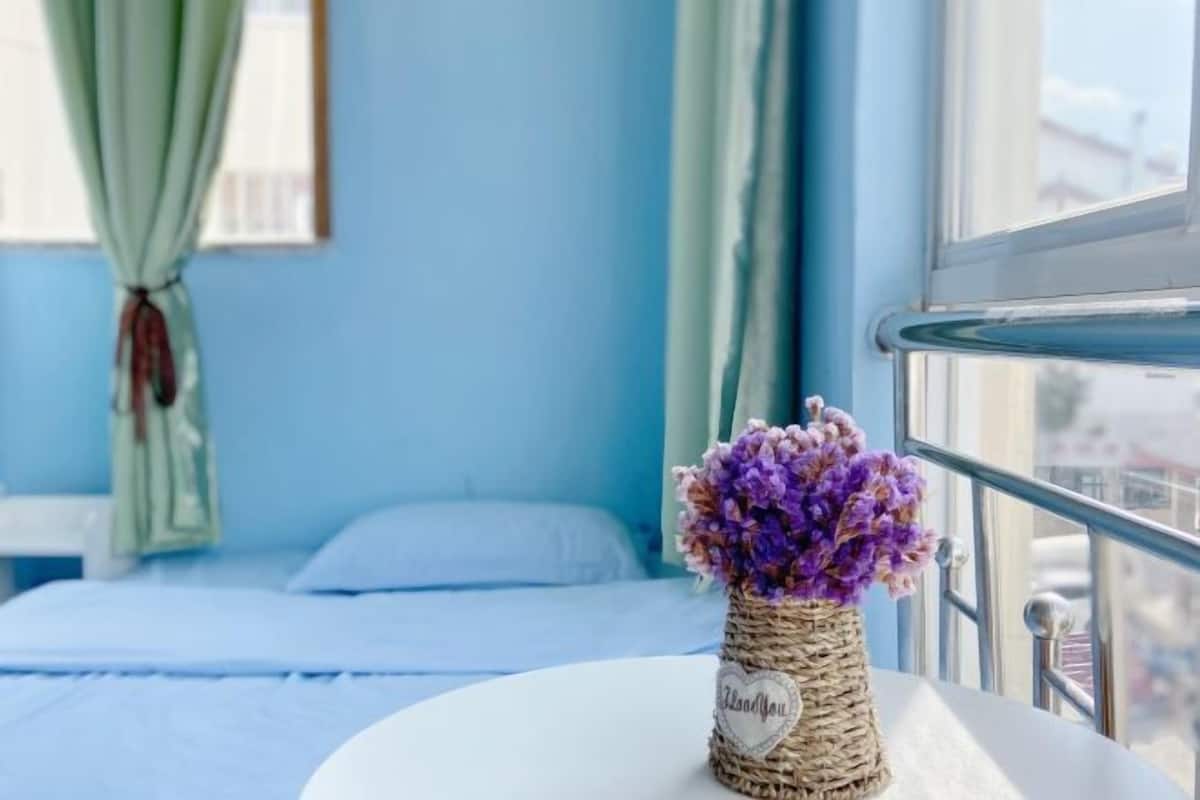 A room with a bed, a window with a view, and a vase with purple flowers on a table.