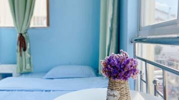 A room with a bed, a window with a view, and a vase with purple flowers on a table.