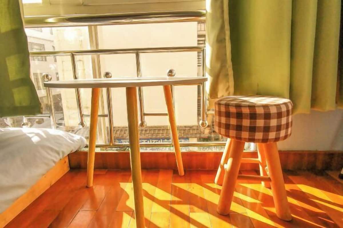 A room with a bed, a table, a checkered stool, and a window with a view of buildings.