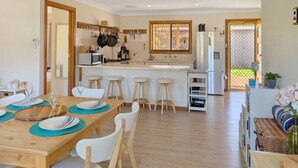 A modern kitchen with a central island, bar stools, and a dining table set for six. - Clifton Cottage by Experience Jervis Bay (Sanctuary Point)