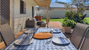An outdoor patio with a table set for four, surrounded by chairs, a wooden serving board, and a small seating area with cushions. - Clifton Cottage by Experience Jervis Bay (Sanctuary Point)