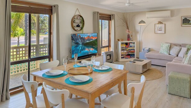 A dining area with a wooden table set for four, a television mounted on the wall, and a bookshelf filled with books. - Clifton Cottage by Experience Jervis Bay (Sanctuary Point)