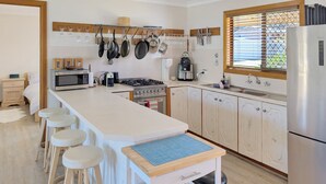 A kitchen with a central island, bar stools, a stove, oven, microwave, and hanging pots. - Clifton Cottage by Experience Jervis Bay (Sanctuary Point)