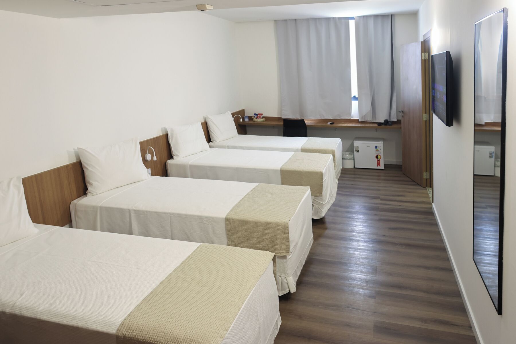 A hotel room with three single beds, a desk, a chair, a small refrigerator, and a television.