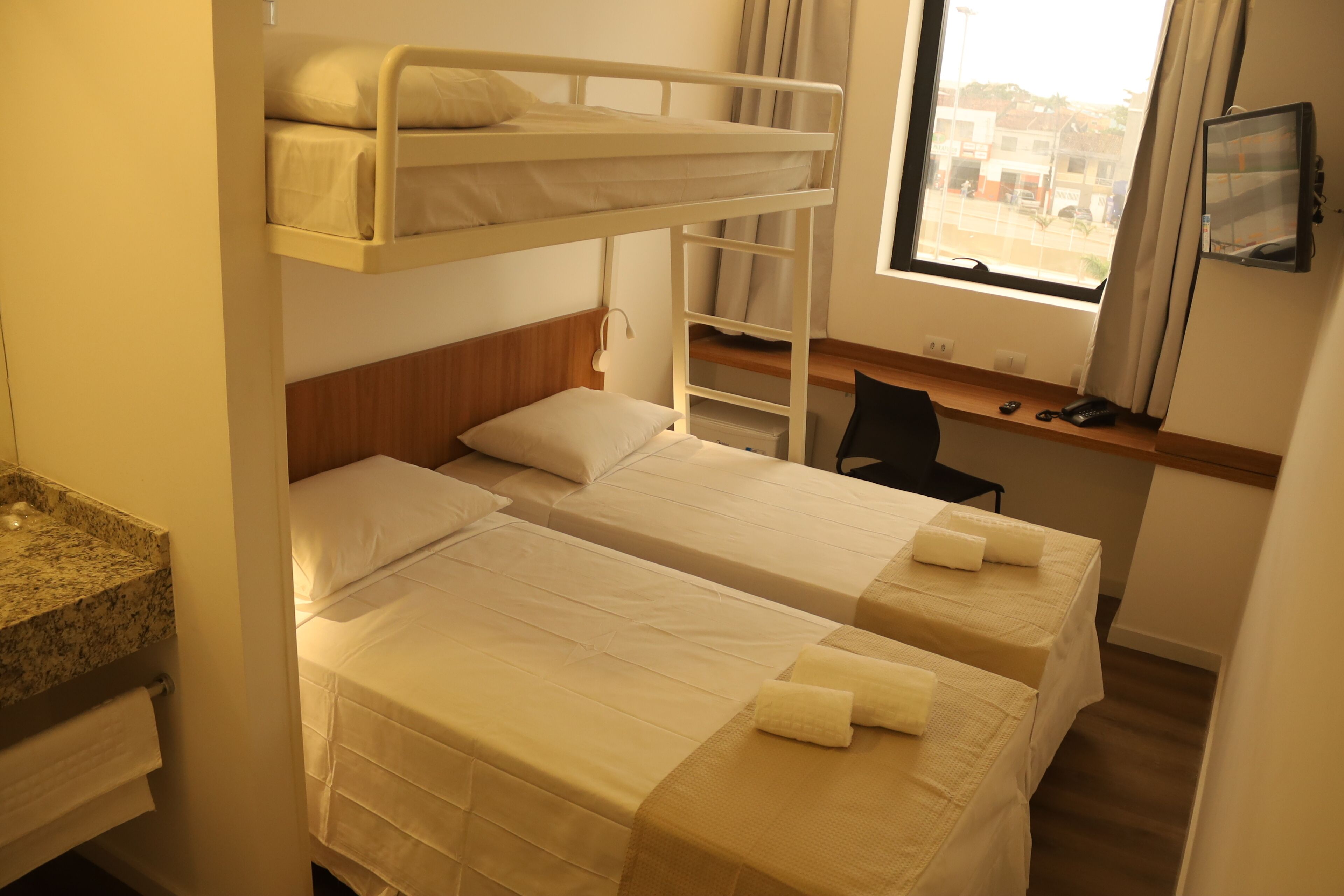Une chambre de dortoir avec des lits superposés, un bureau, une chaise et une fenêtre donnant sur des immeubles.