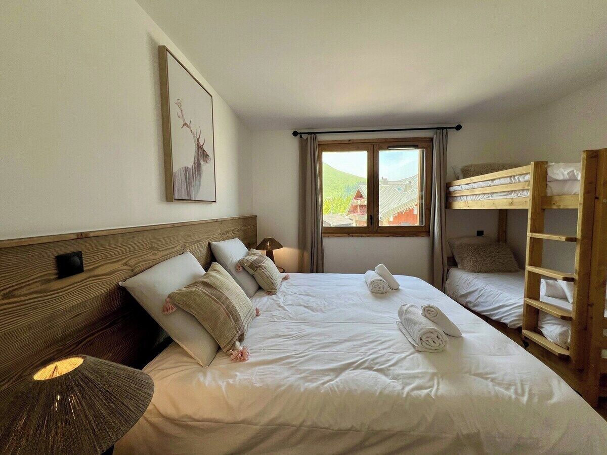 A hotel room with a large bed, a wooden headboard, a bedside table with a lamp, a window with curtains, and a bunk bed in the corner.