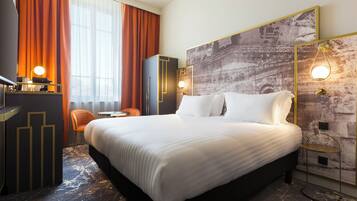 A hotel room with a large bed, a desk with a lamp, a chair, and a wall-mounted TV.