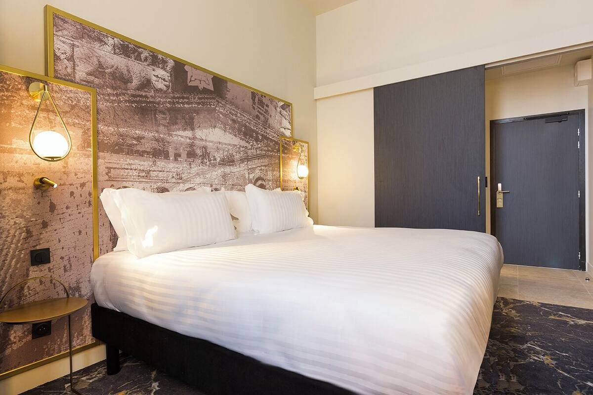 Een hotelkamer met een groot bed, een hoofdbord met een gestructureerde wand, twee bedlampen, een donkere deur en een klein tafeltje.