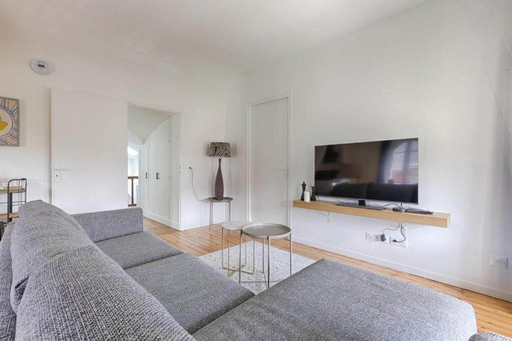 Una sala moderna con un sofá seccional gris, un televisor de pantalla plana montado en un estante de madera y una mesita redonda con una lámpara.