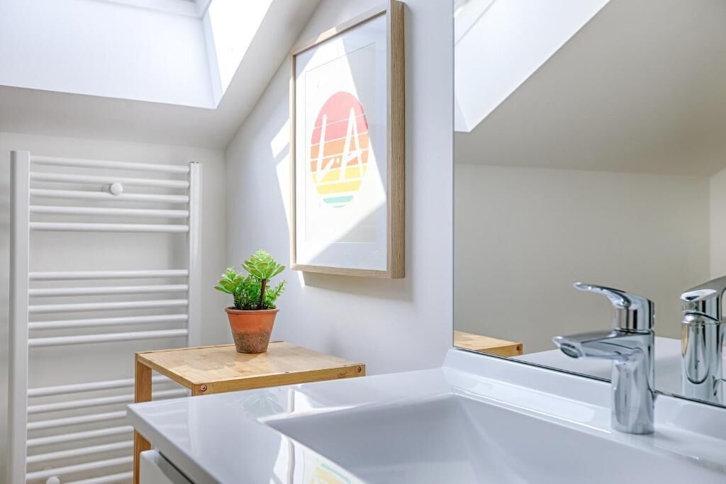 A modern bathroom with a white vanity, a large mirror, a chrome faucet, and a potted plant on a wooden stool.