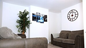 A living room with a brown sofa, a green plant, and a wall-mounted clock. - Cozy Birmingham House - Nec Hs2 Jlr Airport (Birmingham)