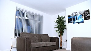 A living room with a brown sofa, a window with blinds, a potted plant, and a wall with a waterfall painting. - Cozy Birmingham House - Nec Hs2 Jlr Airport (Birmingham)