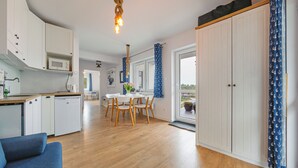 Eine kompakte Küche mit weißen Schränken, einer Mikrowelle und einem Spülbecken. Es gibt einen Essbereich mit Tisch und Stühlen sowie ein großes Fenster mit Vorhängen.