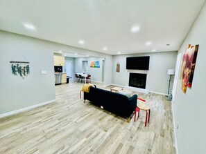 Un salón amplio con un sofá negro, una mesa de centro y una chimenea. Se observa un comedor con mesa y sillas al fondo.
