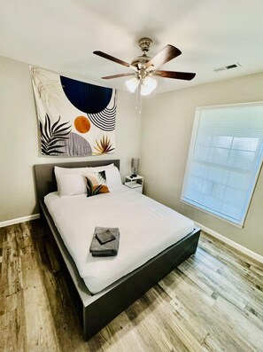Un dormitorio con cama, ventilador de techo, pared con un cuadro abstracto grande y ventana con persianas.
