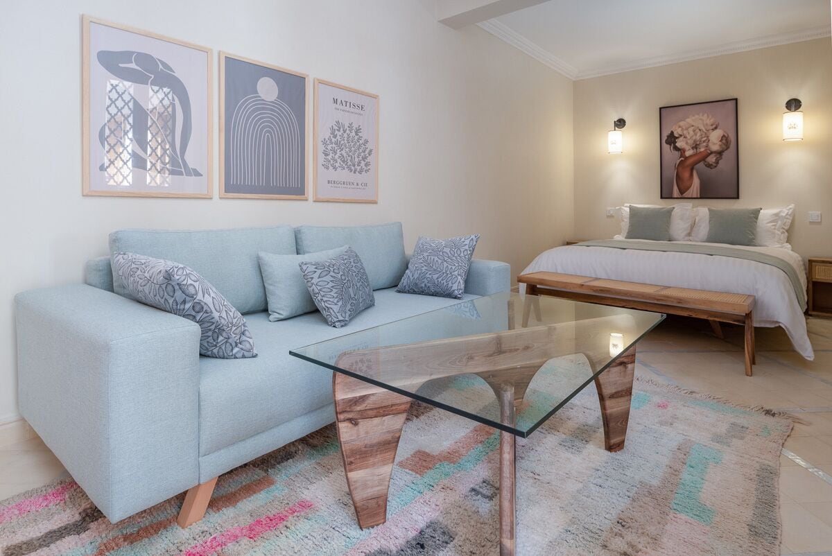Una sala de estar moderna con un sofá azul claro, una mesa de centro de vidrio y una cama con duvet blanco. Hay tres obras de arte enmarcadas en la pared y dos luces fijadas a la pared.