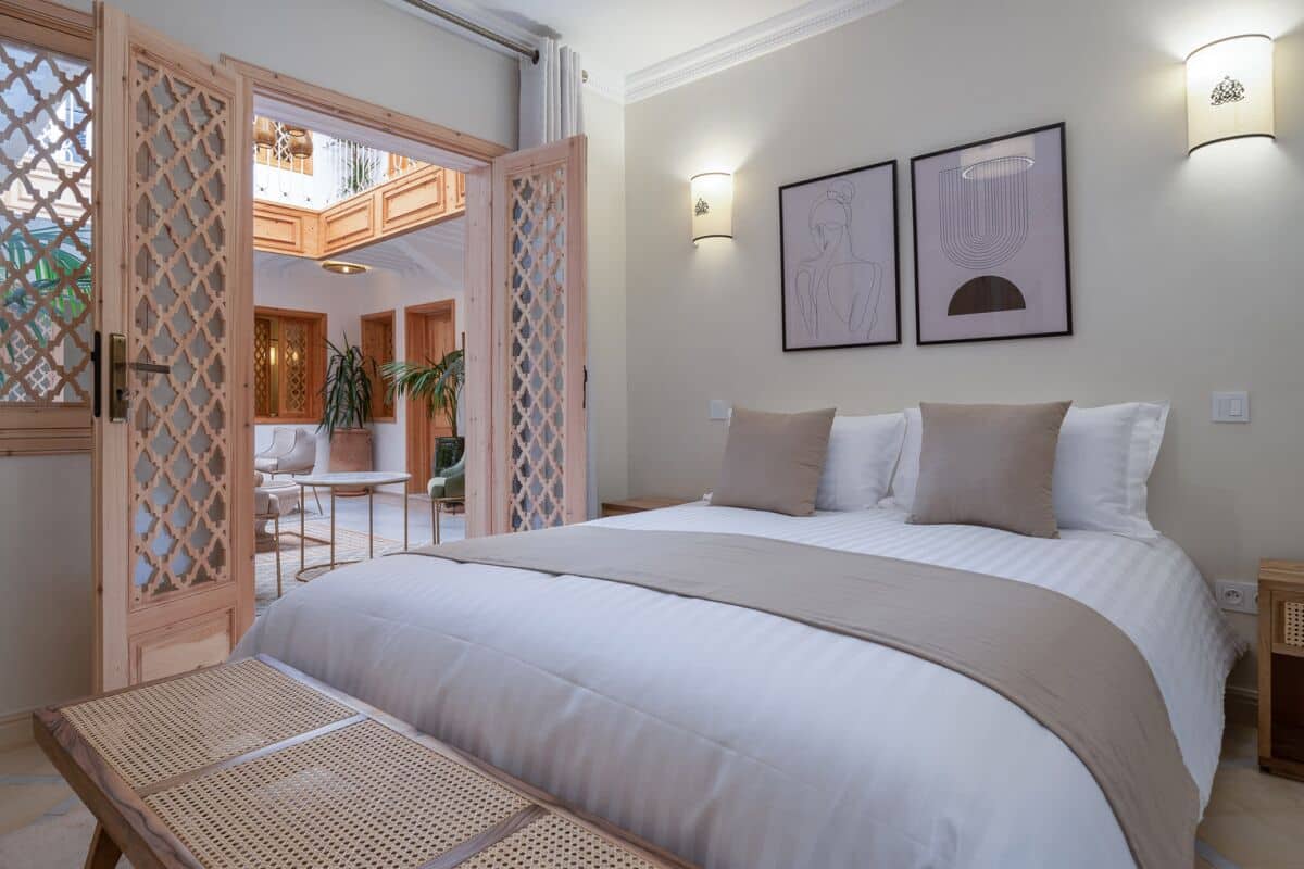 A bedroom with a large bed, a wooden bench, a balcony with a view, and two wall-mounted lights.