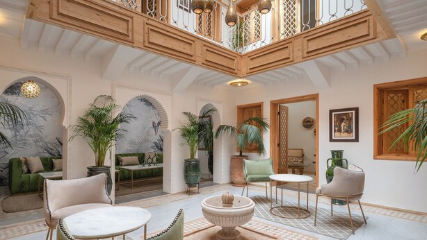 Eine geräumige Lobby mit hoher Decke, Holzbalken und einem Schachbrettmuster-Boden. Es gibt mehrere Sitzbereiche mit Stühlen und Tischen, und um den Raum herum befinden sich Kübelpflanzen.