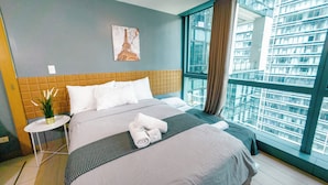 A modern hotel room with a large bed, a small bedside table, a potted plant, and a view of a cityscape through a large window. - Cozi Uptown Executive Suite BGC (Taguig)