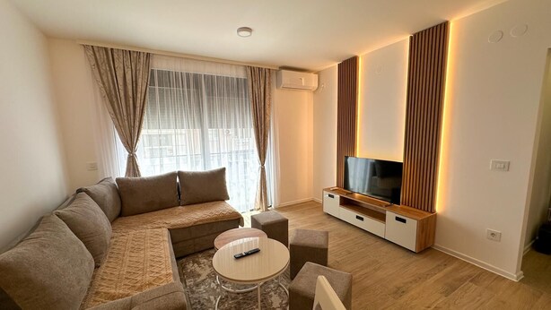 A modern living room with a sectional sofa, a TV on a wooden stand, a small round table, and a window with curtains. - I&I 3 Bedrooms Apartment (Kragujevac)