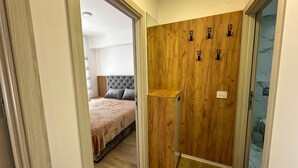 A hallway leading to a bedroom with a wooden wardrobe, a bed with a grey headboard, and a small wooden shelf. - I&I 3 Bedrooms Apartment (Kragujevac)