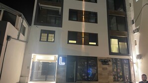 A multi-story building with a garage entrance, a neon 'L' sign, and a car parked outside. - I&I 3 Bedrooms Apartment (Kragujevac)