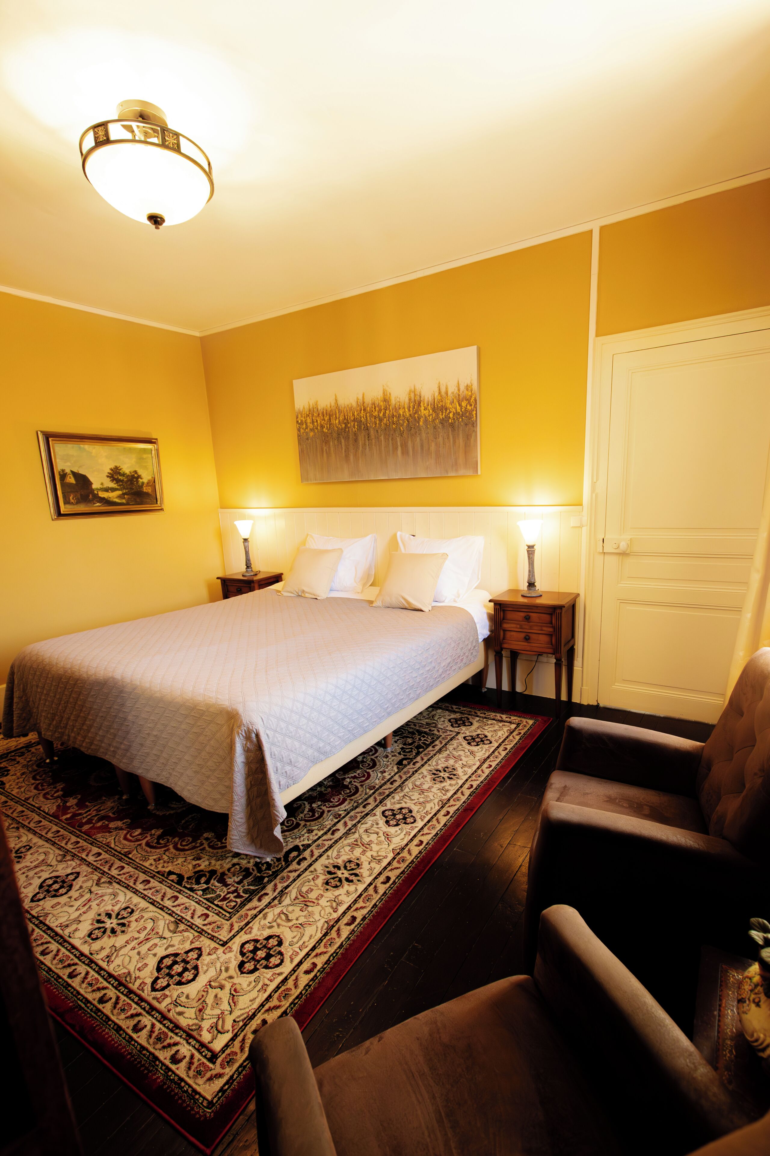 Una habitación de hotel con cama, mesitas de noche, una silla, una puerta y un cuadro en la pared.