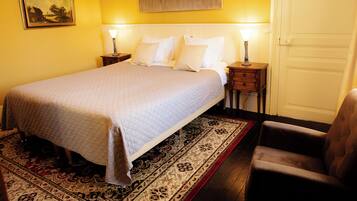 Una habitación de hotel con cama, mesitas de noche, una silla, una puerta y un cuadro en la pared.