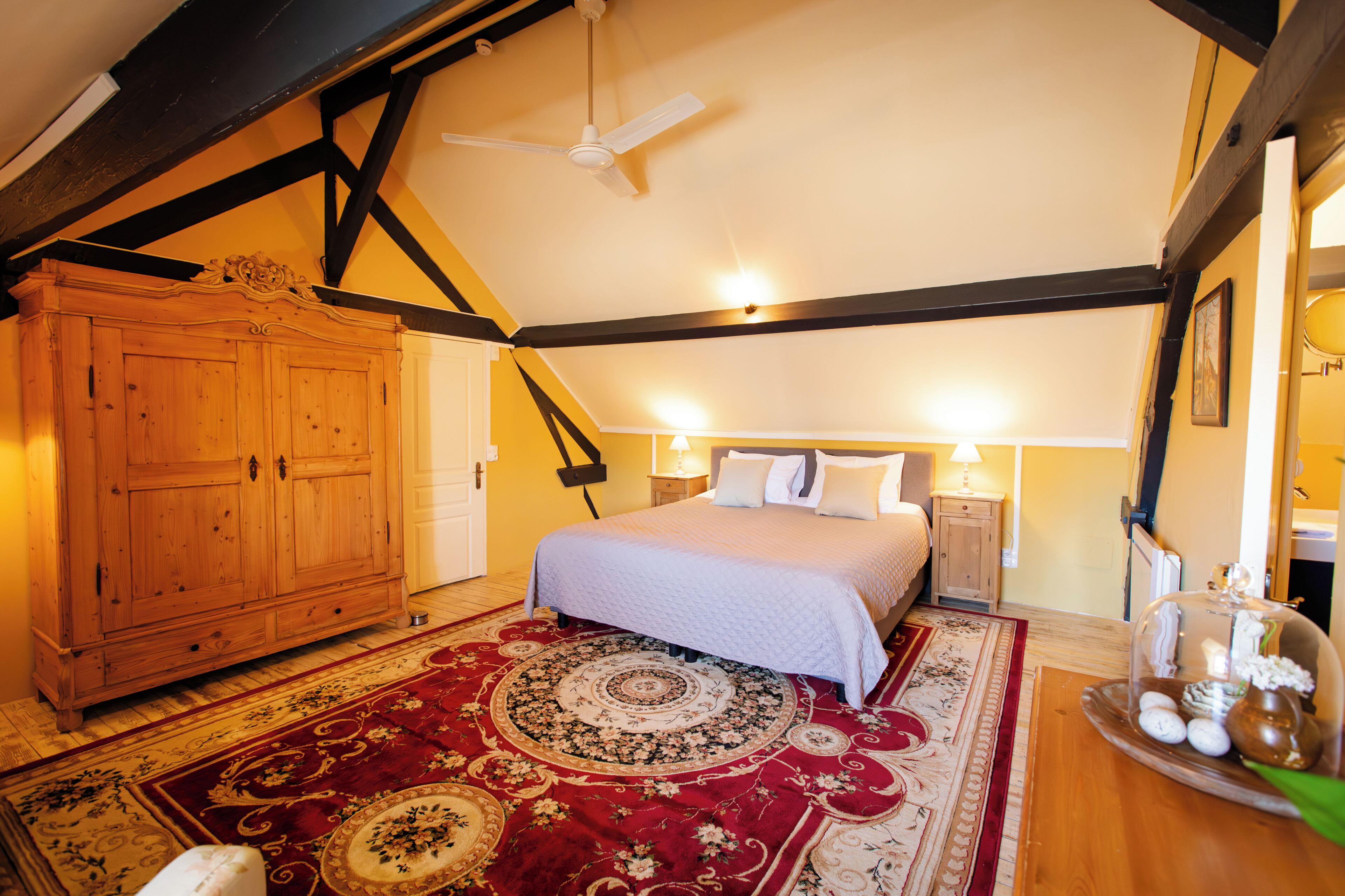 A bedroom with a large bed, a wooden wardrobe, a ceiling fan, and a decorative rug.