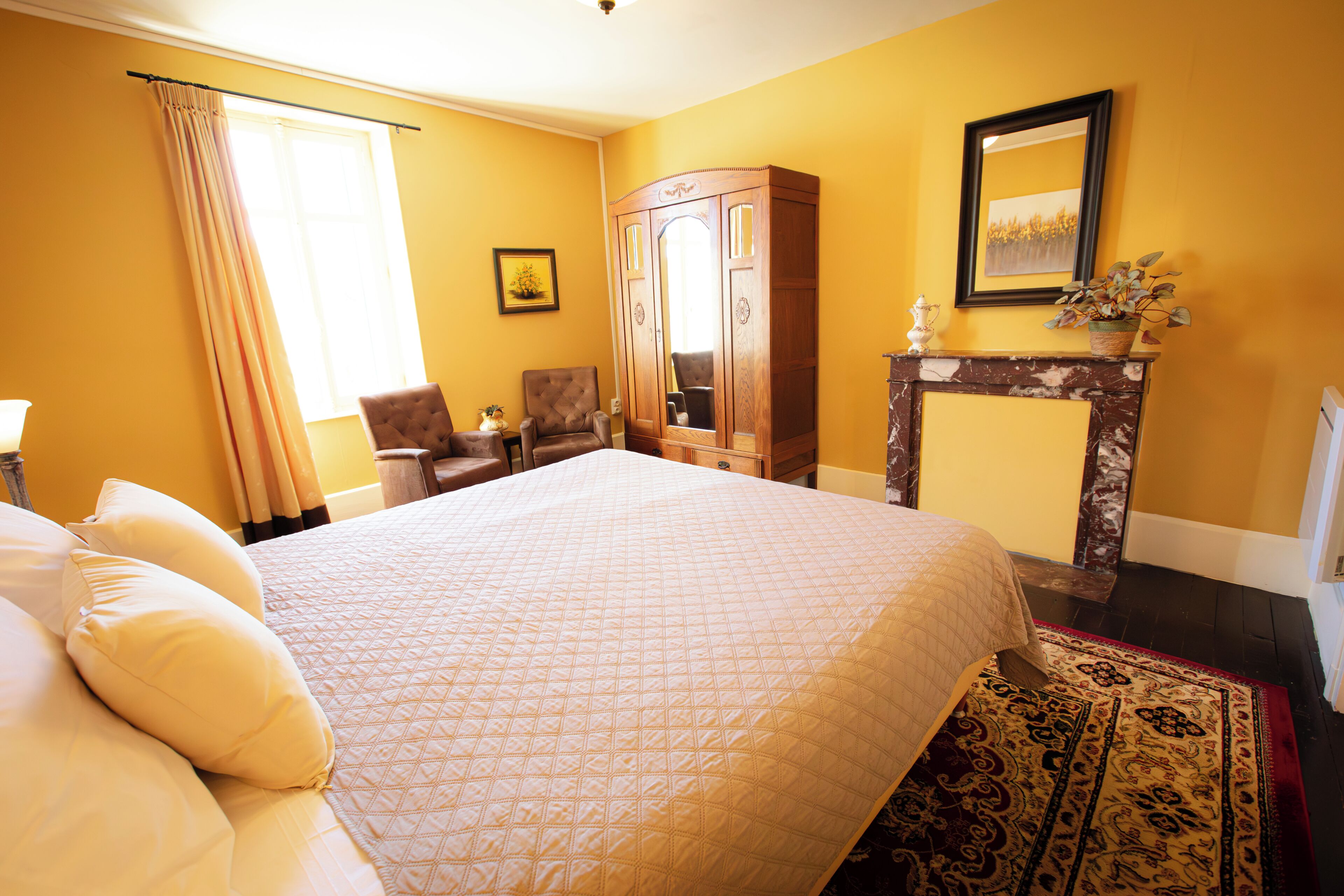 A hotel room with a large bed, a sitting area with a chair, a wooden wardrobe, a fireplace, and a framed picture on the wall.