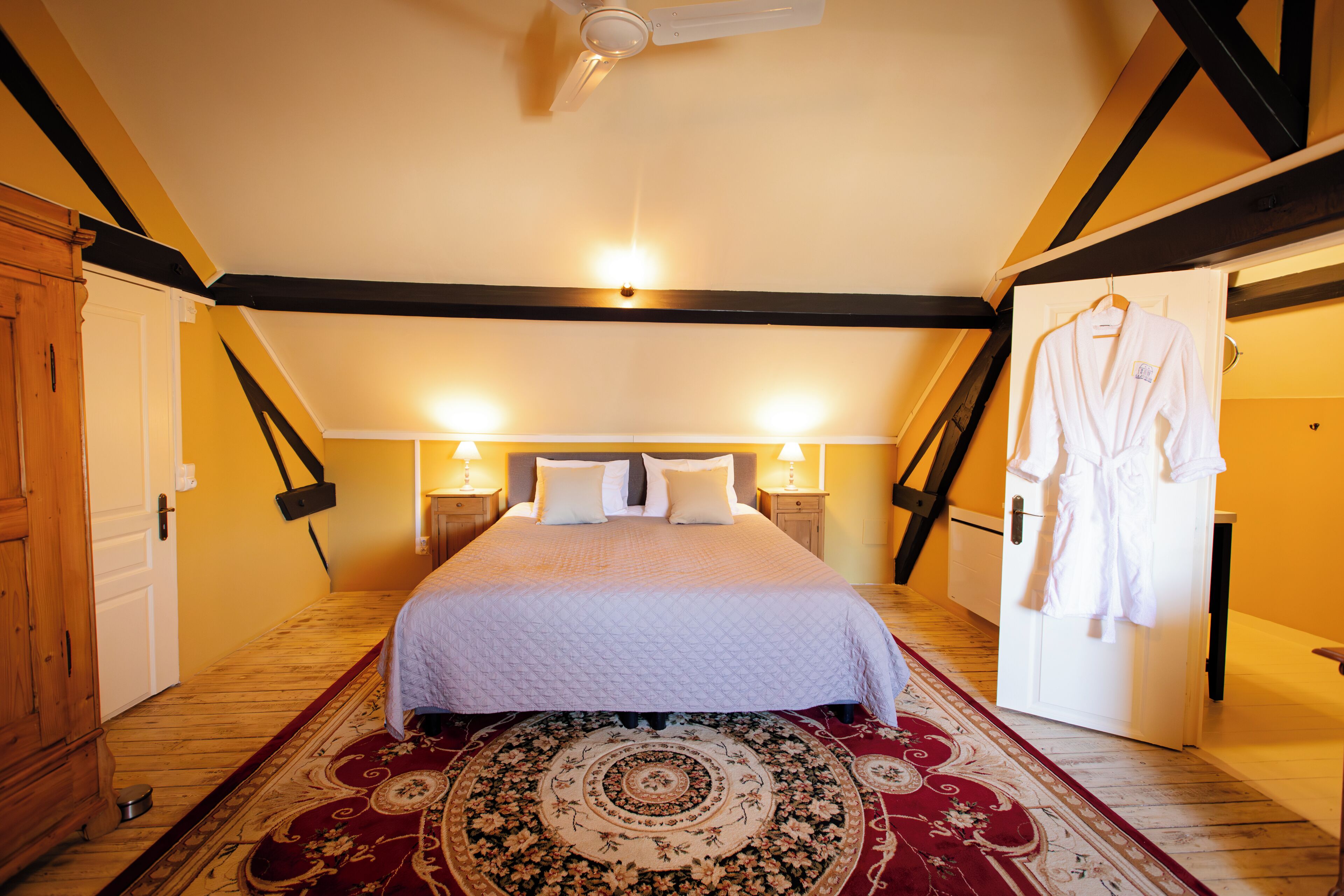 A bedroom with a bed, two bedside lamps, a ceiling fan, a wooden wardrobe, and a robe hanging on the door.