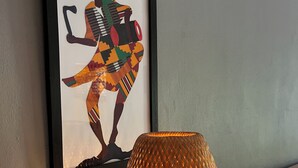 Un'opera d'arte incorniciata raffigura una persona che suona uno strumento musicale tradizionale, una lampada arancione con una superficie strutturata e una testiera di legno.