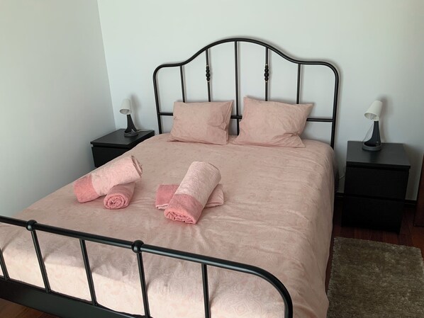 Una cama bien hecha con sábanas y almohadas rosas, dos mesitas de noche con lámparas y suelo de madera.