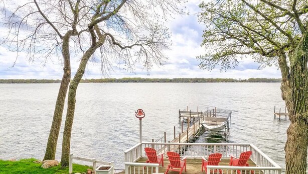 A lakeside deck with red Adirondack chairs, a dock extending into the water, and a white railing. - Colcord Point Sunset Cottage and Guest Cottage (Okoboji)