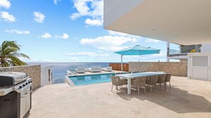 A rooftop pool area with a view of the ocean, a barbecue grill, and a dining table with chairs.