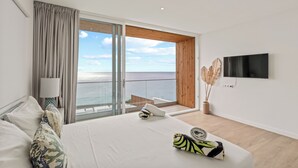 A modern hotel room with a large bed, a TV mounted on the wall, and a balcony with a view of the sea.