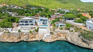 A coastal property with a large house featuring a swimming pool, a rocky shoreline, and a mountainous backdrop.