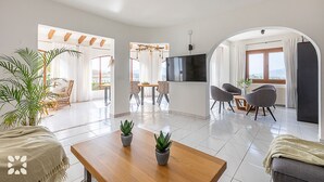 Un vaste espace de vie comprenant une table à manger, des chaises, un téléviseur et une grande fenêtre agrémentée de rideaux.