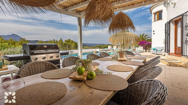 Un espace extérieur aménagé pour les repas, avec un barbecue, des meubles en osier et une vue sur les montagnes.