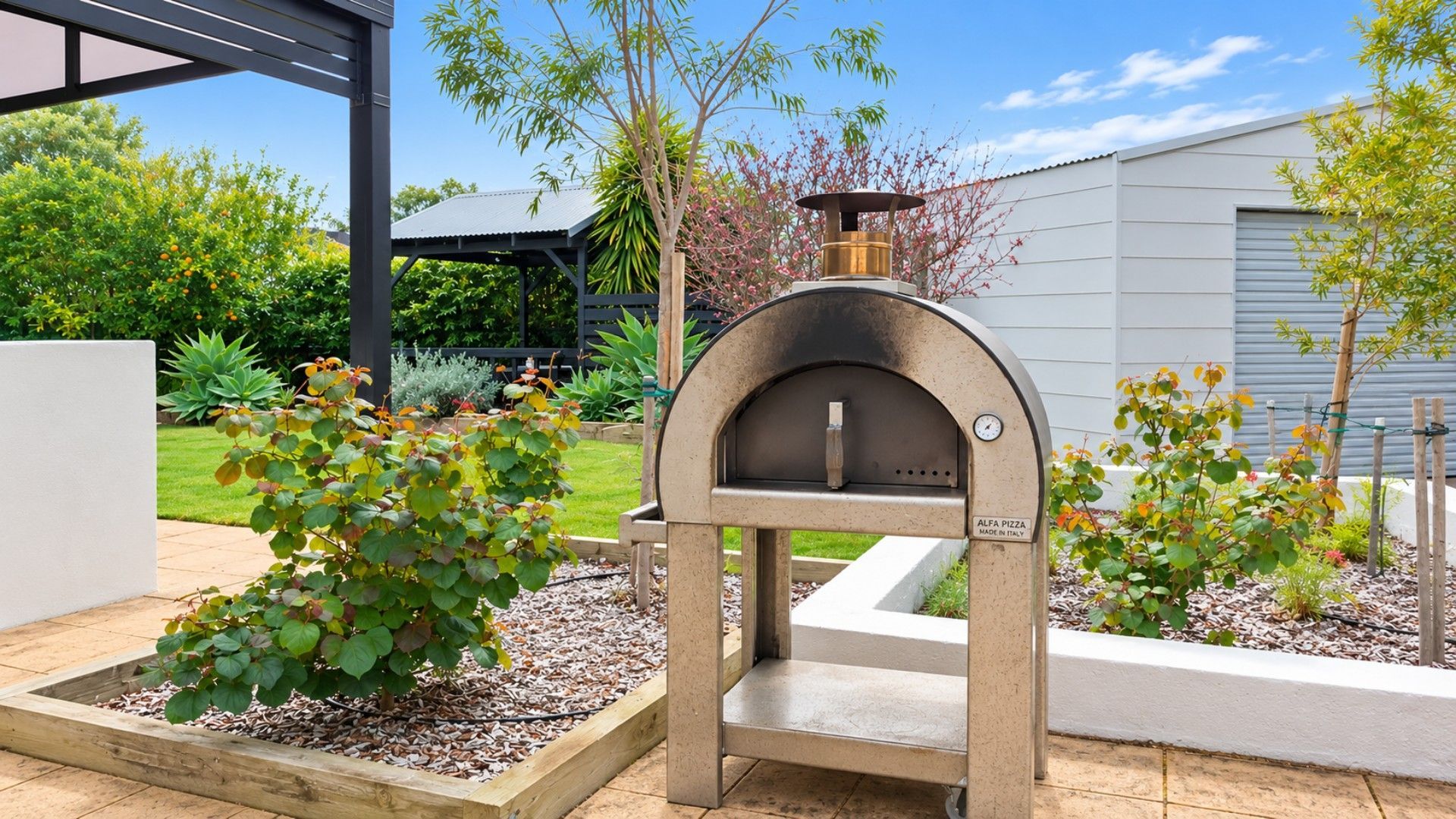 An outdoor pizza oven with a label indicating it is made in Italy, surrounded by a garden with various plants and a modern building in the background.