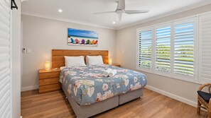 A bedroom with a bed, a nightstand, a lamp, a ceiling fan, a window with blinds, and a painting on the wall.