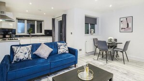 A modern living room with a blue sofa, a dining area with a table and chairs, and a kitchen in the background. - Modern 1BR Flat | Walkable High Street Stay (Southend-on-Sea)